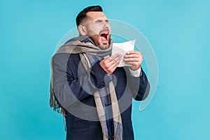 Sick man in official style suit and wrapped in scarf sneezing tissue, feeling unwell with runny nose, suffering allergy or