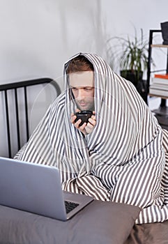 Sick man sitting on bed wrapped in blanked uses laptop