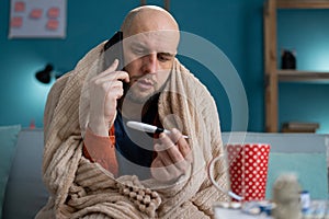 Sick man sitting on sofa in warm blanket, holding thermometer and making phone call to doctor. Flu and fever