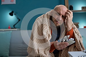 Sick man suffering from fever and headache holds blisters with pills while sitting on the sofa. Treatment and illness
