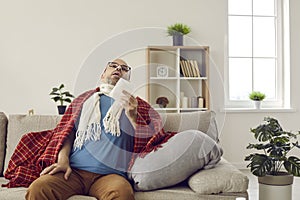 Sick man who has flu or cold sitting on couch at home, holding paper tissue and sneezing