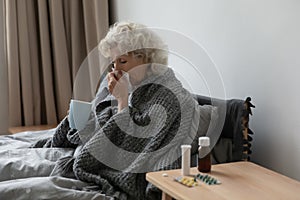 Sick mature woman blowing running nose, sitting in bed