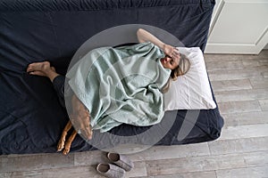 Sick middle age woman at home lying in bed with dog, suffering from headache, flu, touching forehead