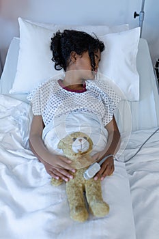 Sick mixed race girl asleep in hospital bed wearing fingertip pulse oximeter and holding teddy bear