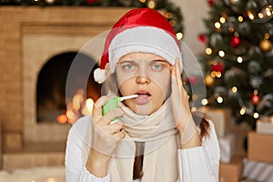 Sick on New Year! Sad ill woman in santa hat and white scarf sitting near Christmas tree and fireplace with inhaler, suffering