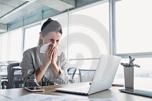 Sick and overworked businesswoman sitting at the desk in the office and blowing her nose, feeling unwell.