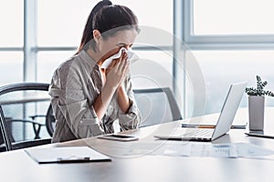 Sick and overworked businesswoman sitting at the desk in the office and blowing her nose, feeling unwell.