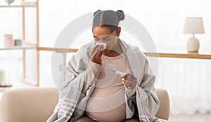 Sick pregnant black woman sneezing and checking body temperature