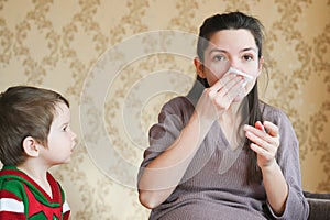 Sick pregnant woman with a runny nose at home. The youngest child is looking at a pregnant mother