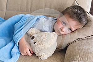 A sick or sad boy lies on a sofa under a blue blanket in an embrace with a teddy bear. Misunderstanding of parents, childhood