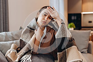 Sick sad girl sitting on the sofa covered with a blanket. He measures the temperature with his hand, shows that he is not well