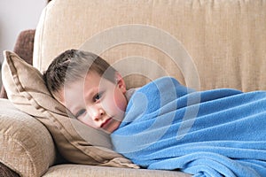 A sick or sad little boy of preschool age lies on a sofa wrapped in a blue plaid. Children`s illnesses, depression, alienation.