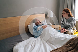 Sick senior aged male patient lying on bed in recovery room at hospital with granddaughter looking after