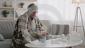 Sick senior man with fever measuring body temperature with thermometer, sitting alone at home interior wrapped in plaid