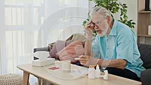 Sick senior man with fever measuring body temperature with thermometer, sitting alone at home interior wrapped in plaid