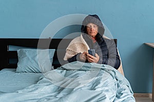 Sick senior man with phone, feel cold covered with blanket sit in bed, shiver freezing warming at home, no central