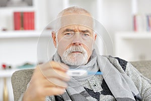 sick senior man in scarf holding thermometer at home