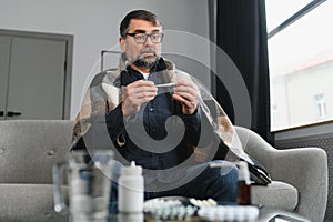 Sick senior man using thermometer checking his temperature suffering from seasonal flu or cold.