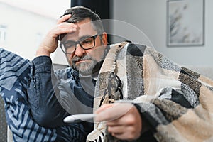 Sick senior man using thermometer checking his temperature suffering from seasonal flu or cold.