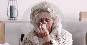 Sick senior woman covered with blanket blowing runny nose