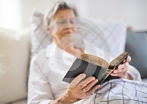 Sick senior woman reading bible book in bed at home or in hospital.