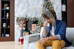 Sick sneezing man with runny nose sitting on sofa at home, hispanic winter day near Christmas tree, cold and allergy in