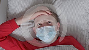 Sick teen boy in a protective mask and red pajamas lies in bed and touches his forehead with his hand. View from above