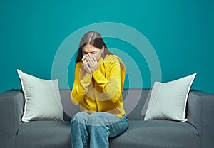 Sick unhealthy allergic young girl blowing running nose, sneezing in tissue, sitting on sofa. Allergy or flu symptoms