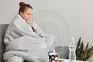 Sick unhealthy woman with hair bun sitting on sofa wrapped in blanket, having illness, suffering runny nose and sneezing, posing