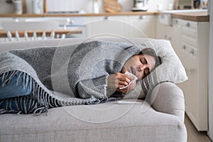 Sick unhealthy woman lying under blanket on sofa coughing and sneezing, using tissue to blow nose