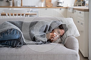 Sick unhealthy woman lying under blanket on sofa coughing and sneezing, using tissue to blow nose