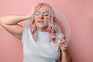 Sick unwell young woman touching head and drinking water while taking painkiller pill against isolated over pink