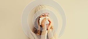 Sick upset woman sneezing blow nose using tissue wearing warm soft knitted clothes, hat and sweater on beige studio background