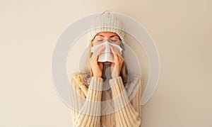 Sick upset woman sneezing blow nose using tissue wearing warm soft knitted clothes, hat and sweater on beige studio background
