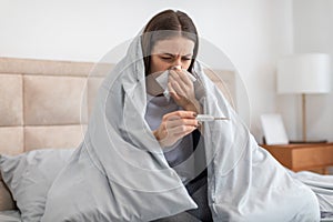 Sick woman in bed, checking temperature with thermometer, feeling unwell