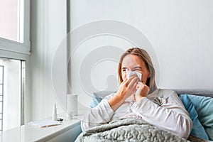 Sick woman blowing her nose with headache and fever lying under the blanket. Sick woman staying in bed with temperature durong