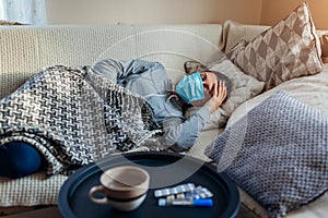 Sick woman having flu or cold. Girl lying in bed wearing protective mask by pills and water on table