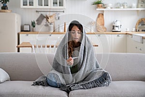 Sick woman sitting on sofa covered blanket looking at electronic thermometer measuring temperature