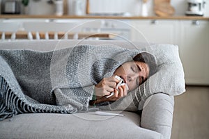 Sick woman with weakened immunity sneezing in tissue while lying under blanket on sofa at home
