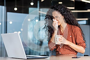 Sick woman working in office, hispanic woman having flu and cold drinking hot tea sneezing and coughing at workplace