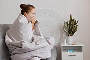 Sick young adult woman with hair bun sitting on sofa wrapped in blanket, having symptoms of pneumonia and bronchitis, coughing,