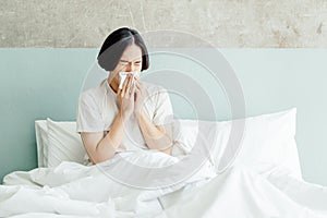 Sick young Asian man blowing his nose while sitting on bed at home