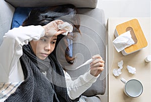 Sick young Asian woman feeling cold lying down on sofa at home. Girl measuring her temperature with a thermometer and hand