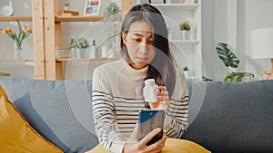 Sick young Asian woman hold medicine sit on couch video call with phone consult with doctor at home. Girl take medicine after