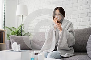 Sick young asian woman sit under blanket on sofa. Female blow nose, coughing, sneezing, check temp in tissue at home, suffering