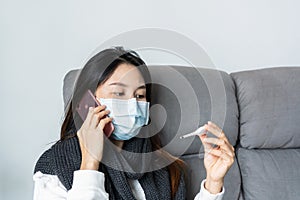 Sick young Asian woman sitting on sofa at home while looks at thermometer and talking on phone with doctor for health counseling