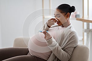 Sick young black expecting woman sneezing and checking temperature