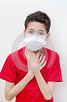 Sick young boy clutches his throat with face mask on.