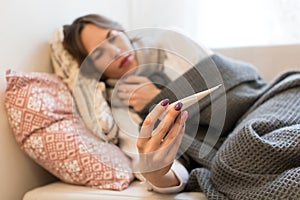 Sick woman lying on sofa under the coverlet, having flu symptoms, looking at thermometer.Virus