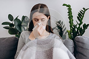 Sick young caucasian woman sitting on grey sofa in living room freezing blowing running nose got fever caught cold sneezing in